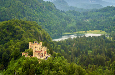 A view of a Hoehenschwangau castle surrounded by forest and a Swan lake in the background.