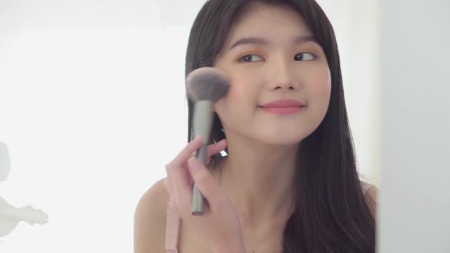 Beauty young asian woman happy and smile with face looking mirror applying makeup with brusher of cheek in the room, beautiful of girl holding brush make-up on facial, skin care and cosmetic concept.