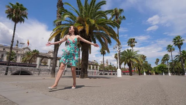 Happy tourist woman run and whirl around at Moll de Bosch i Alsina, pedestrian alley against Port Vell in Barcelona. Empty passage, tall palm trees grow around. Excited lady spin and jog