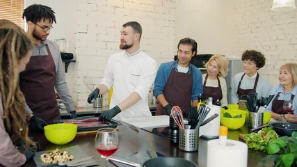 Diverse group of people in aprons are cooking food during cookery courses in kitchen discussing culinary with chef in uniform. Meals and occupation concept.