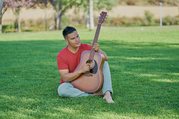Hispanic male musician playing acoustic guitar standing on green grass and barefoot in a cheerful attitude