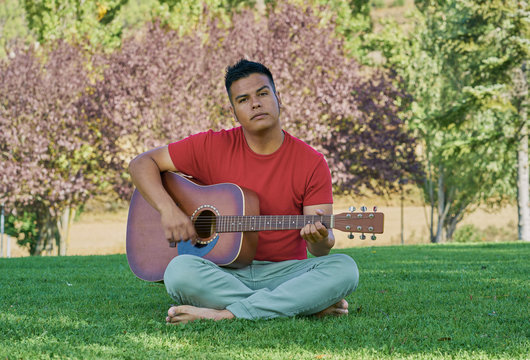 Hispanic male musician playing acoustic guitar standing on green grass and barefoot in a cheerful attitude
