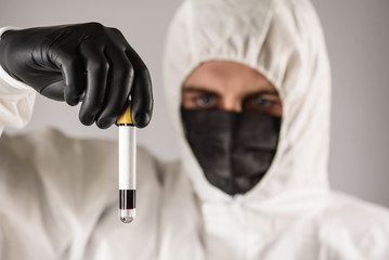 a virologist in a black mask and medical gloves wearing a white protective suit, conducts blood...