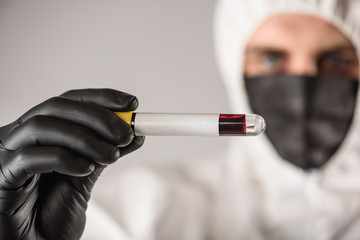 a virologist in a black mask and medical gloves wearing a white protective suit, conducts blood...