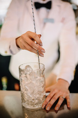 Female bartender in a white suite and black bow tie mixing cocktail in pitcher using long bartenders spoon. Smooth image with shallow depth of field.