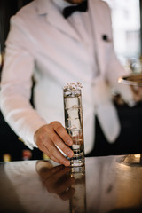 Bartender in a white suite and black bow tie holding on his hand gin and tonic cocktail in a highball glass decorated with a bunch of dried flowers. Smooth image with shallow depth of field.