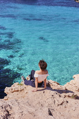Sunny sea day. Young female blogger sitting on the rock above the sea and looking away. Young woman traveler enjoying beautiful view and dreaming about something