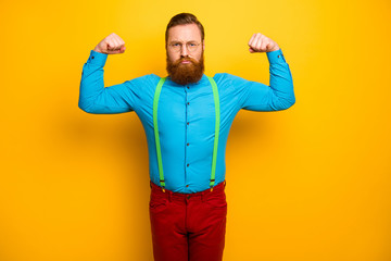 Photo of attractive funny guy seriously raise fists arms presenting perfect shape biceps sports addicted wear blue shirt green suspenders red pants isolated yellow color background - obrazy, fototapety, plakaty