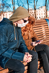 Fototapeta premium Young man and woman wearing the face masks to avoid virus infection and to prevent the spread of disease. Couple sitting on a bench in city centre. Virus infection protection