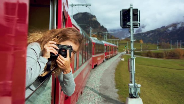 Beautiful happy young woman hangs out of train open window and make photo on vintage analog camera. Travel and explore more wanderlust concept. Female touristin switzerland alp mountains