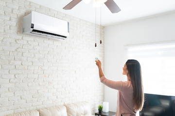 Woman Adjusting Temperature Of Air Conditioner With Remote - obrazy, fototapety, plakaty