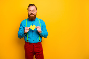 Photo of handsome funny guy hold little paper heart postcard showing feelings look curious empty space wear blue shirt green suspenders red pants isolated yellow color background