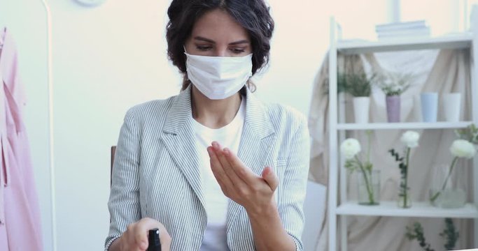 Young businesswoman wears medical face mask using antibacterial sanitizer spray rubbing hands sits at home office desk. Female employee doing personal hygiene disinfection for coronavirus protection.