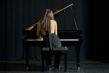 A woman playing a grand piano on a stage. Back view with black dress