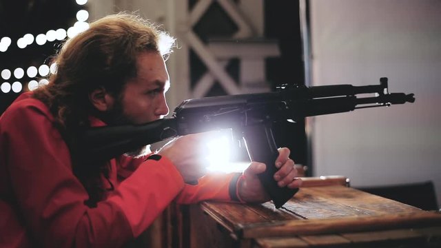A blond-haired and bearded caucasian millennial man shoots from a machine gun in a shooting range late at night, male persone holds weapon in his hands, takes aim, leans on box, lights in background.