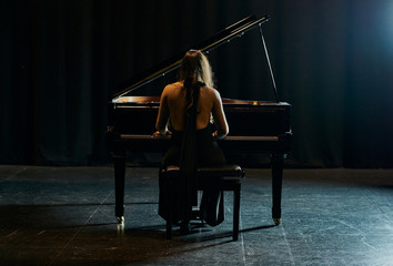 A woman from behind dressed in a black dress playing a black grand piano with the lid raised on a stage © Sergio