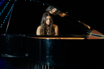 A woman playing a grand piano dressed in a black dress and her face is reflected in the open piano...