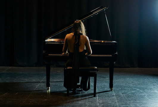 A woman from behind dressed in a black dress playing a black grand piano with the lid raised on a stage
