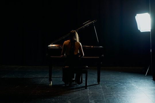 A Woman From Behind Dressed In A Black Dress Playing A Black Grand Piano With The Lid Raised On A Stage