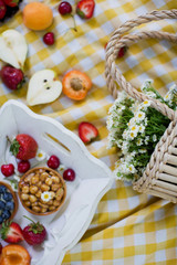 Picnic at the park. Fresh fruits and tarts on a white wooden tray, straw bag with white chamomiles