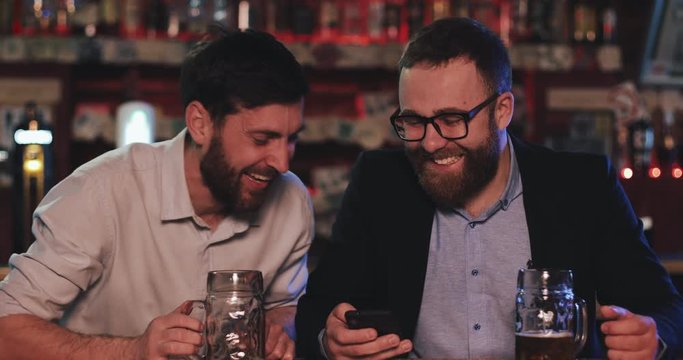 Cheerful men wearing formal suits on night out drinking draft beer on counter. Berded guy in glasses showing funny photo on smartphone to his friend and laughing while sitting at pub.