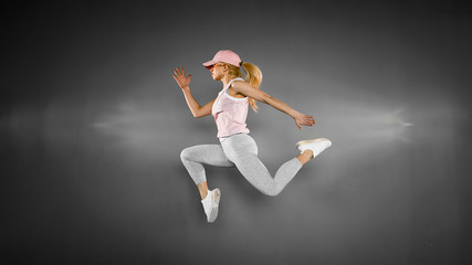 Fototapeta na wymiar Young blonde woman with fit body jumping and running against grey background. Female model in sportswear exercising.