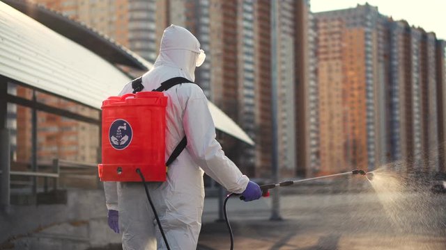 A man in protective equipment disinfects with a sprayer in the city. Surface treatment due to coronavirus covid-19 disease. A man in a white suit disinfects the street with a spray gun. Virus pandemic