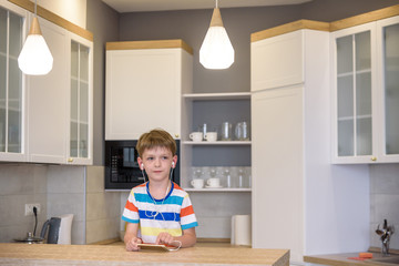 Little boy sitting at kitchen table holding a phone in his hands with earphones while looking to screen, messaging online using Wi-Fi having astonished expression