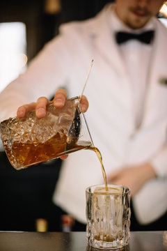 Bartender in a white suite and black bow tie pouring a brown color cocktail from a mixing pitcher into a rocks glass with large ice cube using strainer. Smooth image with shallow depth of field.