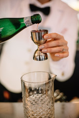 Female bartender in a white suite and black bow tie pouring a pink color cordial into jigger. Smooth image with shallow depth of field.