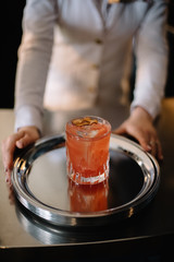 Female bartender holding on her hands metal tray with peach color cocktail in a rocks glass decorated with a dried slice of peach. Dark background. Smooth image with shallow depth of field.