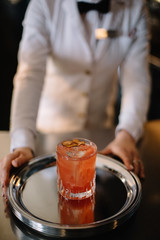 Female bartender holding on her hands metal tray with peach color cocktail in a rocks glass decorated with a dried slice of peach. Dark background. Smooth image with shallow depth of field.