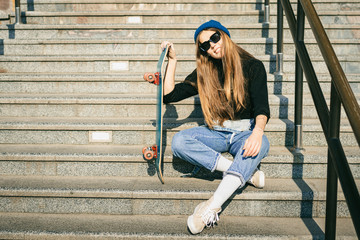 Stylishly dressed woman in blue denim jumpsuit posing with skateboard. Street photo. Portrait of...