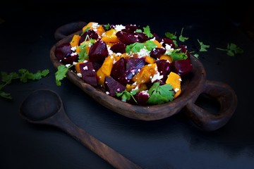 Beetroot and butternut salad with feta and corriander.  Colorful beetroot and butternut cubes on a wooden bowl decorated with crumbled feta and coriander leaves against a black background.