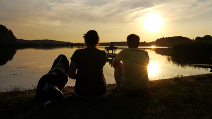 Rodzinne wakacje nad jeziorem z psem. Mężczyzna i kobieta siedzą na trawie nad wodą z psem rasy Basset Hound. Patrzą na zachód słońca nad jeziorem. Przyjaciele, para, zakochani, romantyczny widok. - obrazy, fototapety, plakaty