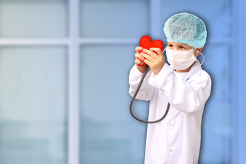 child in a white doctor’s coat, hat and mask attached a stethoscope to a red heart model,...