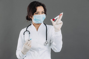 Female doctor with stethoscope in mask holding injection, antidote against coronavirus on gray...