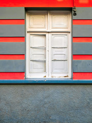 A simple closed window with white shutters in a red and gray wall.