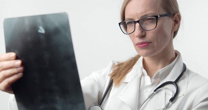 Portrait of mature lady in medical clothing and eyeglasses examining x-ray picture, isolated over white studio background. Concept of diagnosis and pneumonia
