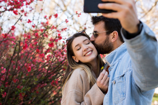 Man and woman taking a cute selfie outside, while the man is kissing the womans cheek.