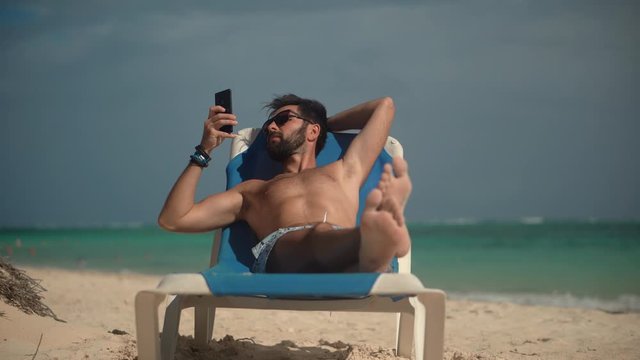 Man Using Mobile Phone On Vacation.Traveller Looking At Cell Phone Using App And Browsing In Internet.Handsome Guy Scrolling On Smartphone Texting Messages On Social Networks And Lying On  Sunbed.