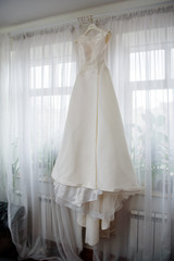 Elegant wedding white dress hanging on a window during a wedding preparation. Bride's morning. Before ceremony