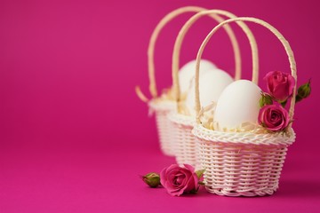 Fototapeta na wymiar White chicken eggs in a white wicker basket covered with straw and small pink roses on a bright pink background. Holiday card with space for text.Easter symbol.Copy space.