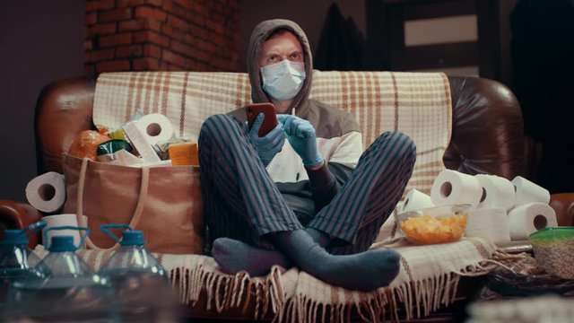 Scared caucasian man in medical mask follows pandemic covid-19 news on internet from smartphone and TV. Quarantine, self-isolation at home during coronavirus epidemic