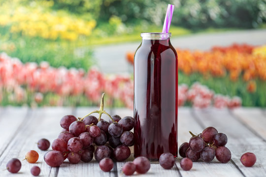 A close up view of a glass bottle of grape juice surrounded by grapes against a garden background.