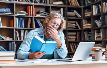 Bored tired sleepy guy university student yawning reading book studying in library feel lack of...