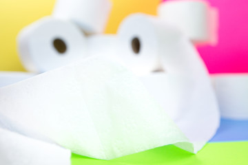 Blurred pile of toilet paper rolls and one roll in front with paper go inco camera making road on yellow blue pink color background hoarding amid panic buying for corona virus outbreak shortage.