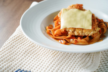 Pasta, Italian food, with tomato sauce topping with deep fried crispy pork cutlet and a sheet of cheese in white dish place on a cloth and on dark brown wooden table and blue plastic utensil near it