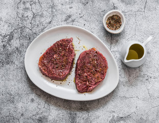 Raw marinated beef steak, olive oil and pepper on a grey background, top view. Food ingredients