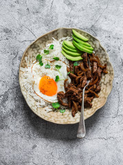 Spicy fried beef, rice, fried egg and avocado - delicious asian style lunch on a grey background, top view